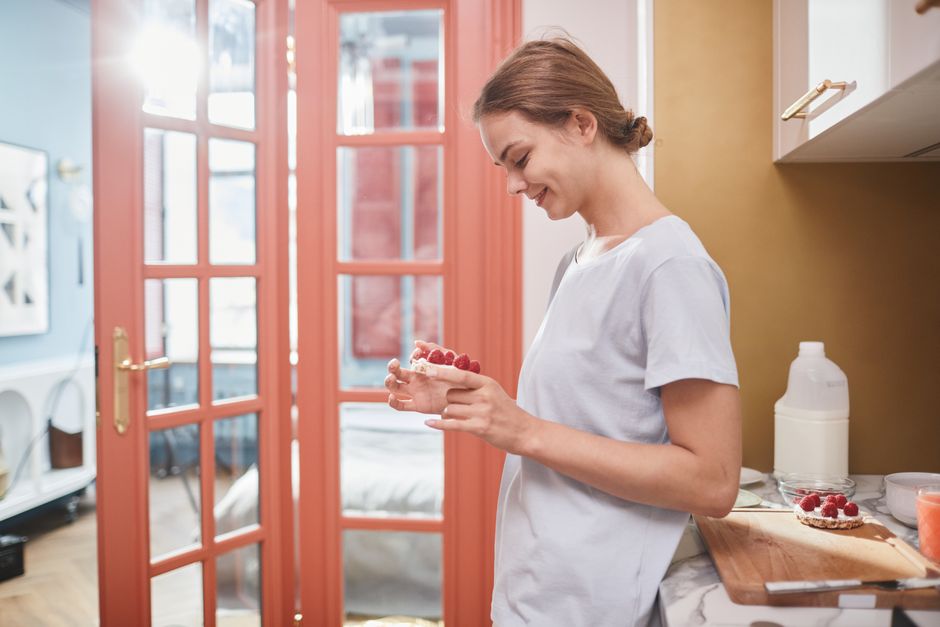 Smiling woman in kitchen holding a healthy breakfast with raspberries, enjoying her morning routine.